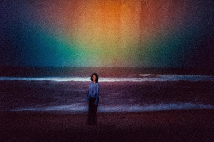 A woman stands on a beach with waves crashing behind her, under a sky with a rainbow-like light effect.