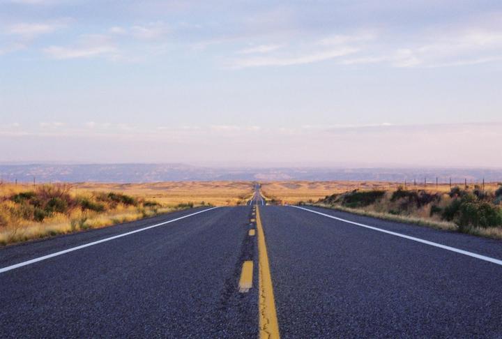 A long, straight road stretches into the distance through a dry, grassy landscape under a pale sky.
