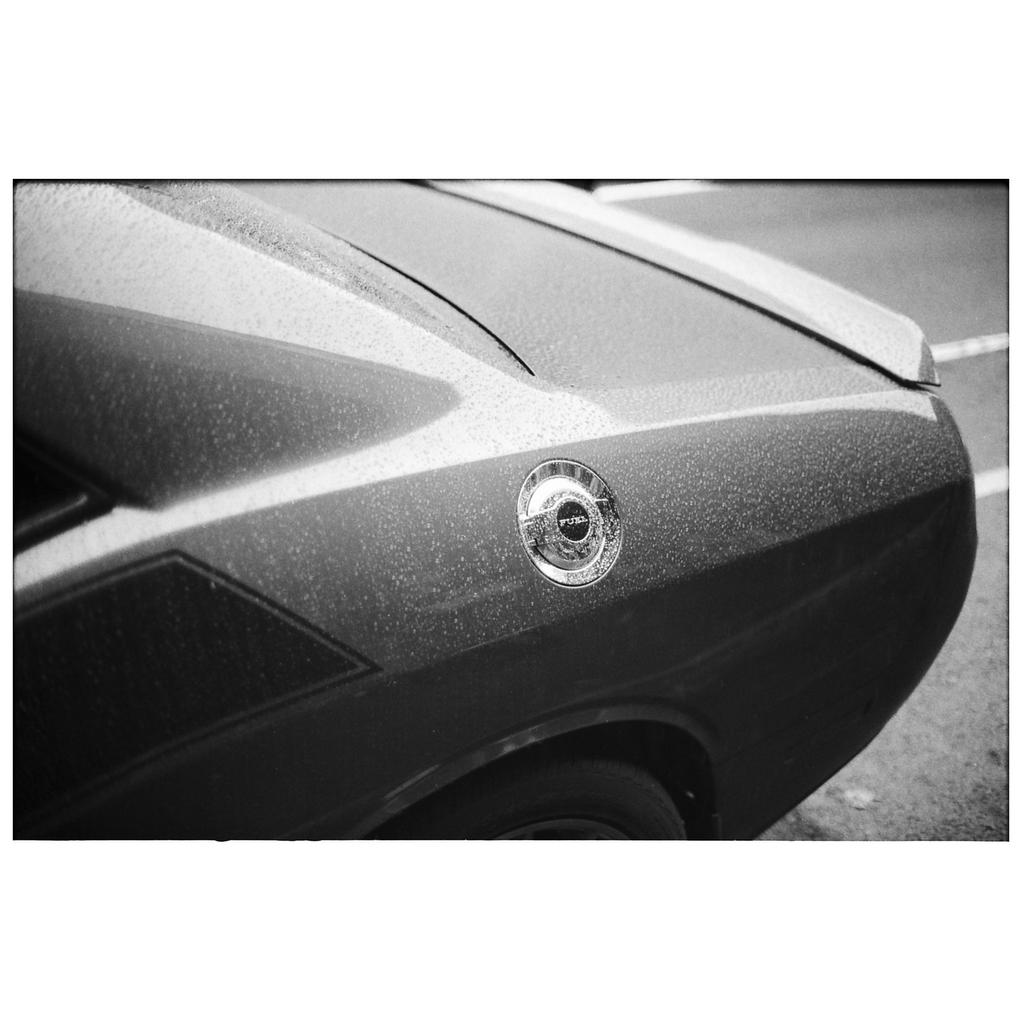 The rear quarter panel of a car with a fuel cap is covered in raindrops.