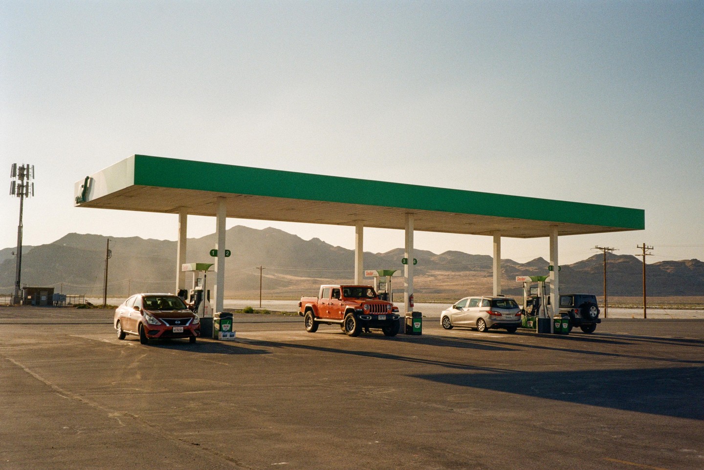 Several vehicles are parked at a gas station with a green canopy in a desert landscape with mountains in the background.