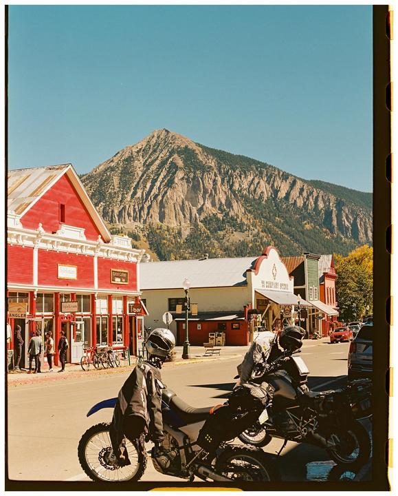 Two motorcycles are parked on a street in front of red buildings with a large mountain in the background.