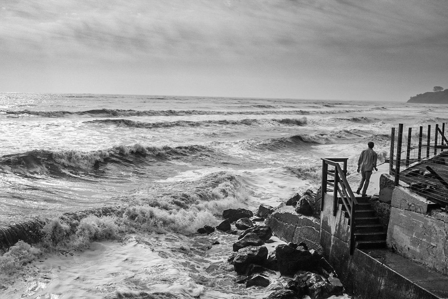 A man walks down concrete steps towards a rough sea with waves crashing against rocks.