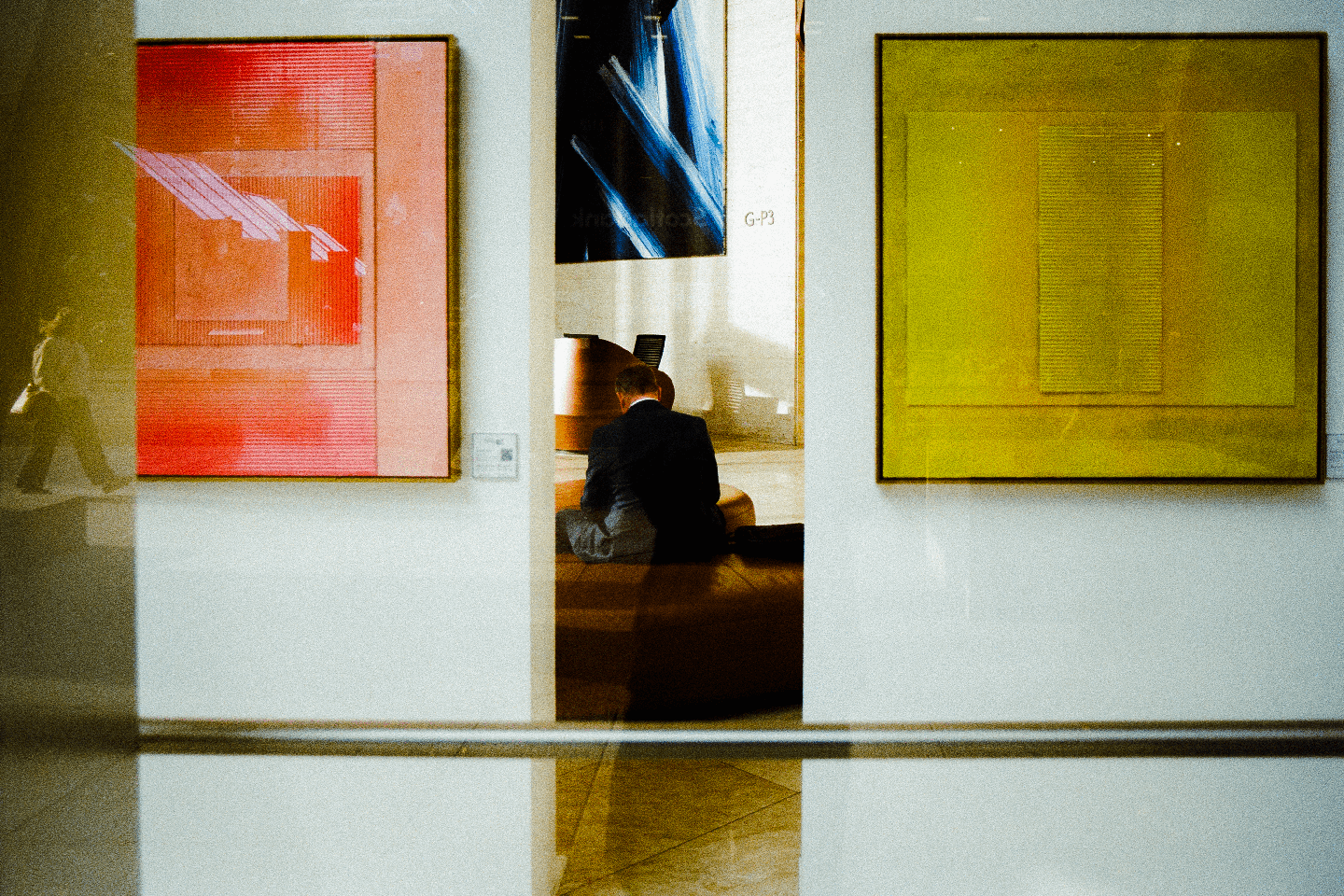 A man sits on a bench between two large abstract paintings, with a person walking past on the left.