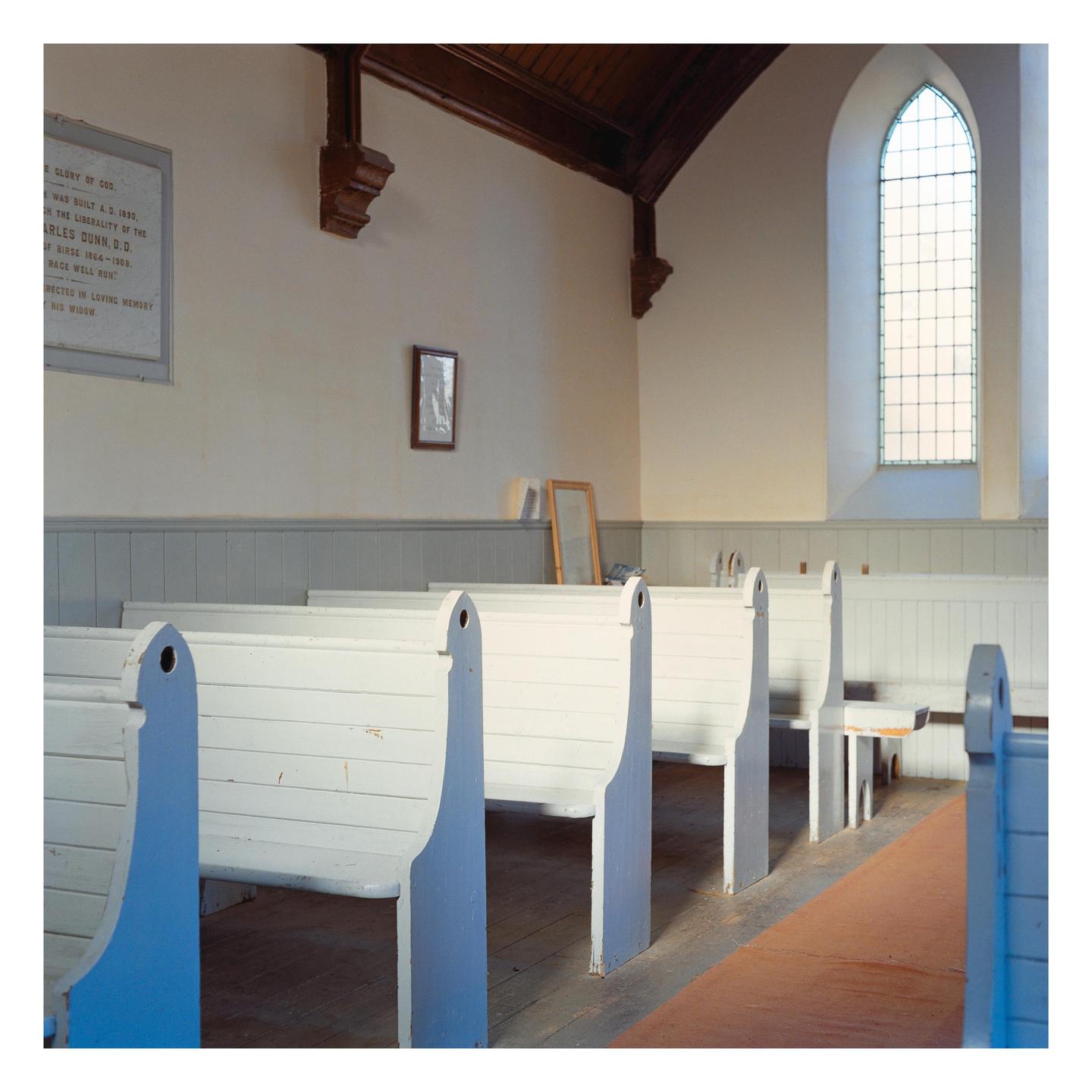 White wooden pews line a church interior with a tall arched window and a wooden ceiling.