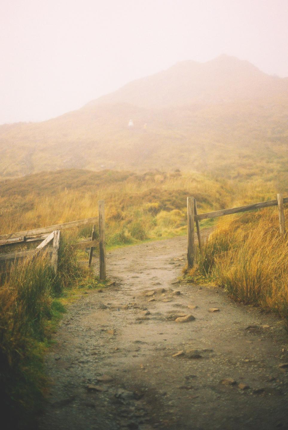 A dirt path leads through a wooden fence towards a hazy mountain.