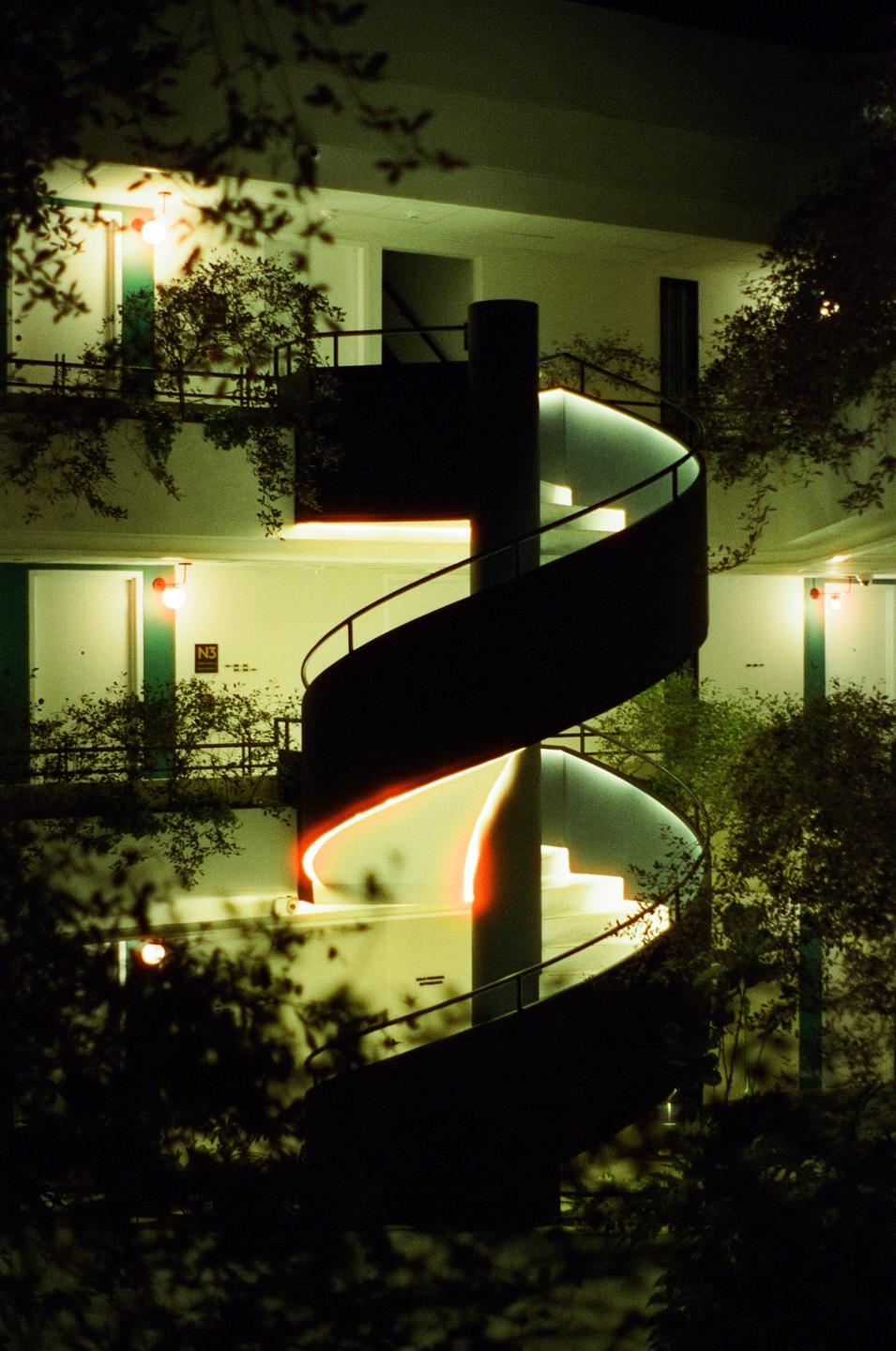 A spiral staircase with glowing orange and white lights is surrounded by green foliage at night.