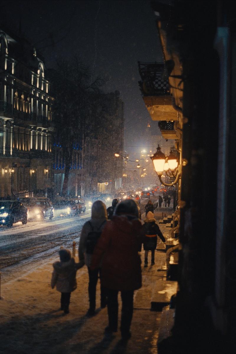 People walk on a snowy street at night, with cars driving by and buildings lit up in the background.