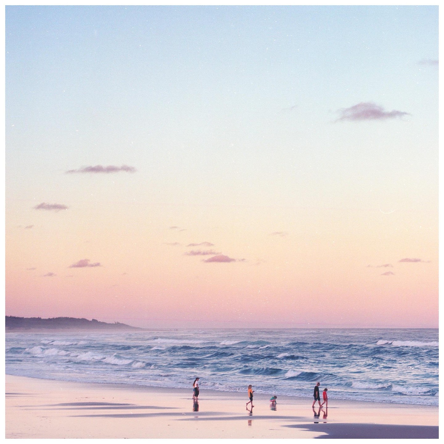 People walk along a beach at sunset with waves crashing on the shore and a headland in the distance.