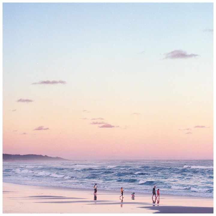 People walk along a beach at sunset with waves crashing on the shore and a headland in the distance.
