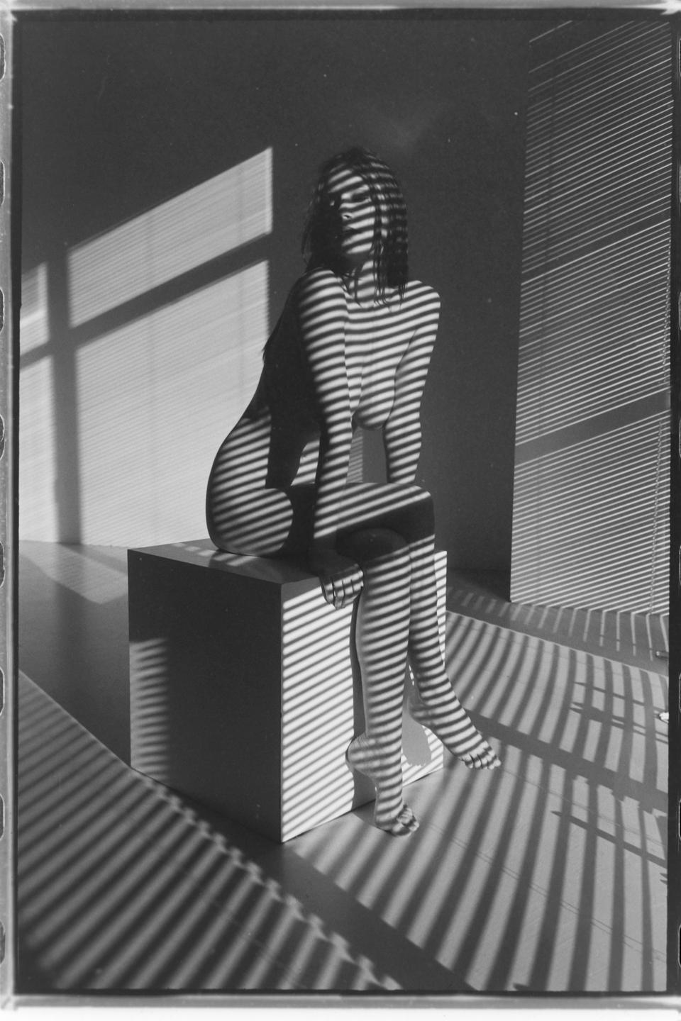 A woman sits on a cube with shadow stripes from window blinds covering her body and the room.