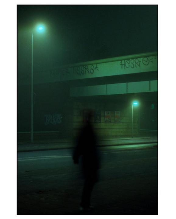 A blurry figure stands on a street at night under a bridge with graffiti, illuminated by streetlights.