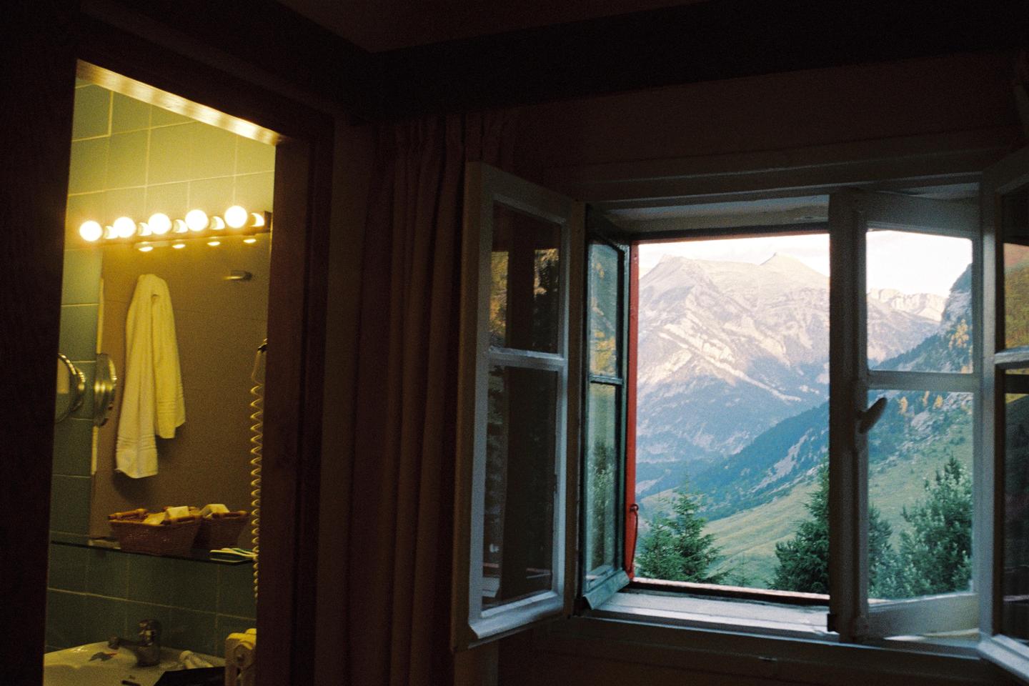 A bathroom with a towel hanging on the wall is visible through a doorway next to an open window looking out at a mountain landscape.
