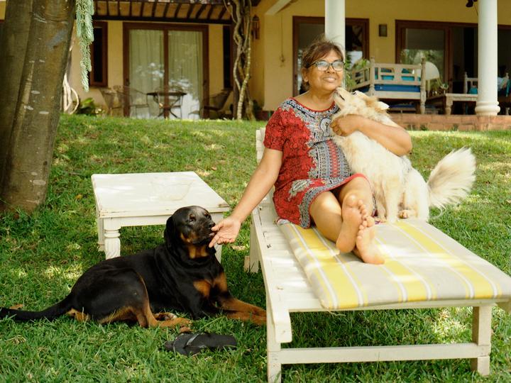 A woman sits on a lounge chair with a fluffy dog in her lap while another dog lies on the grass and licks her hand.