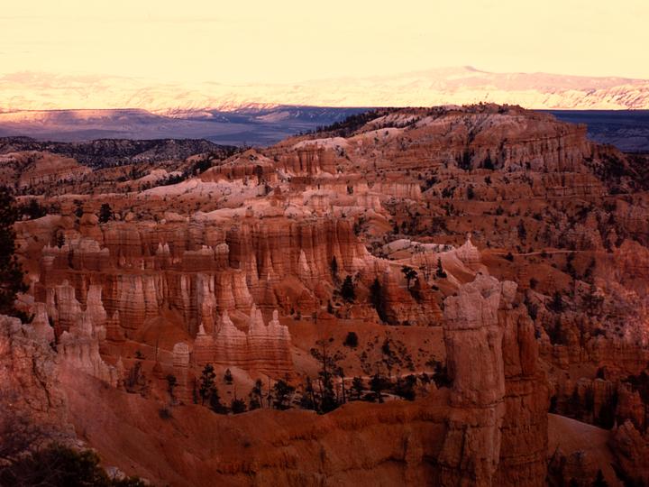 A vast landscape of red rock formations and distant mountains is illuminated by the setting sun.