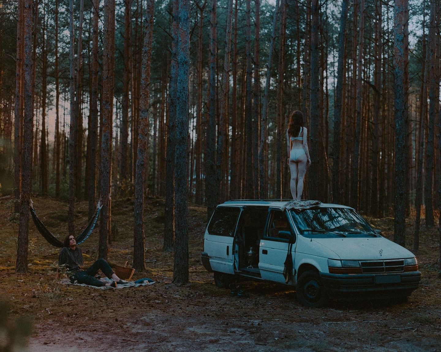 A woman stands on top of a white van in a forest while a man relaxes on a blanket nearby.