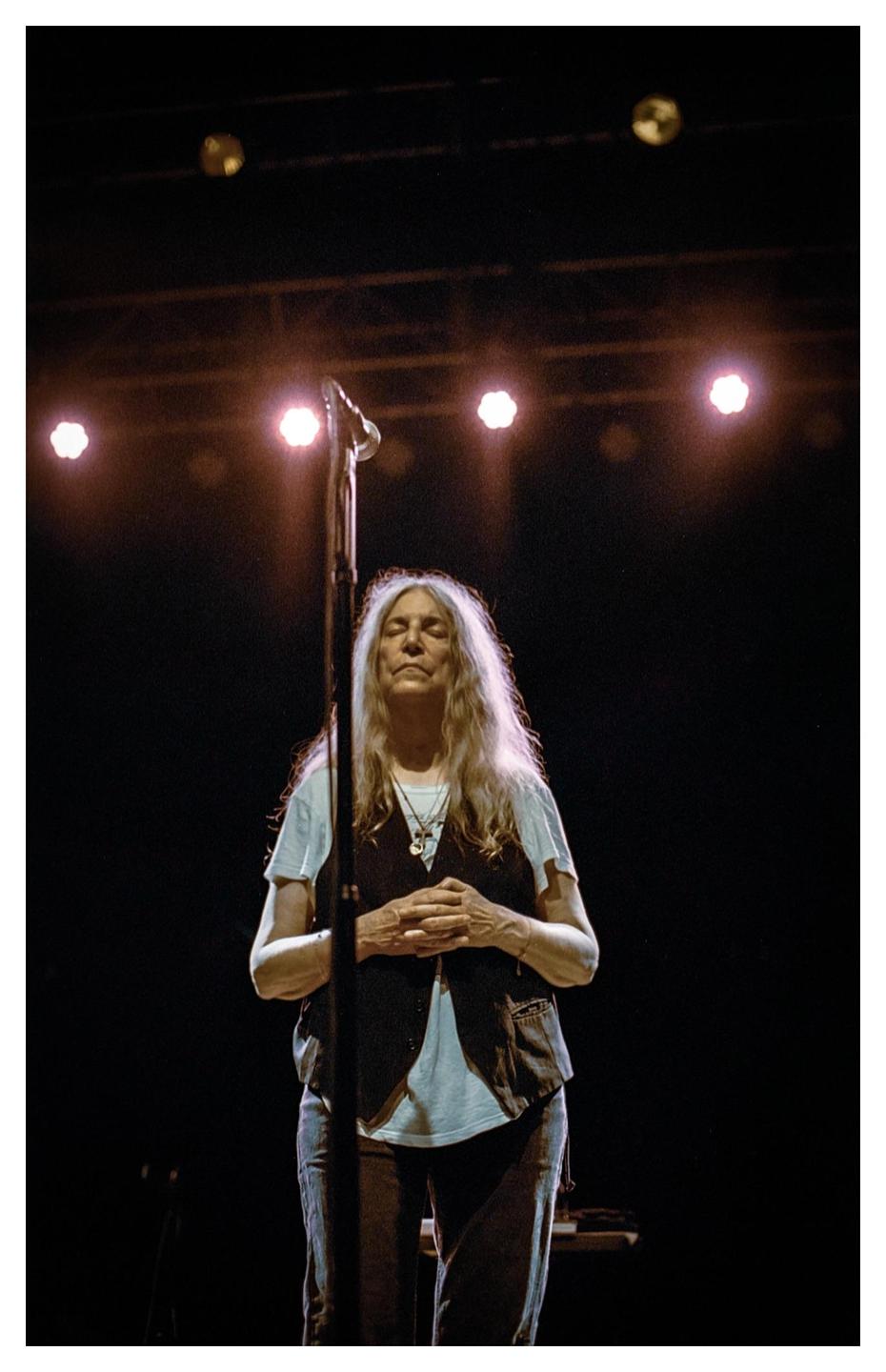 A woman with long hair stands on a stage with her eyes closed and hands clasped.