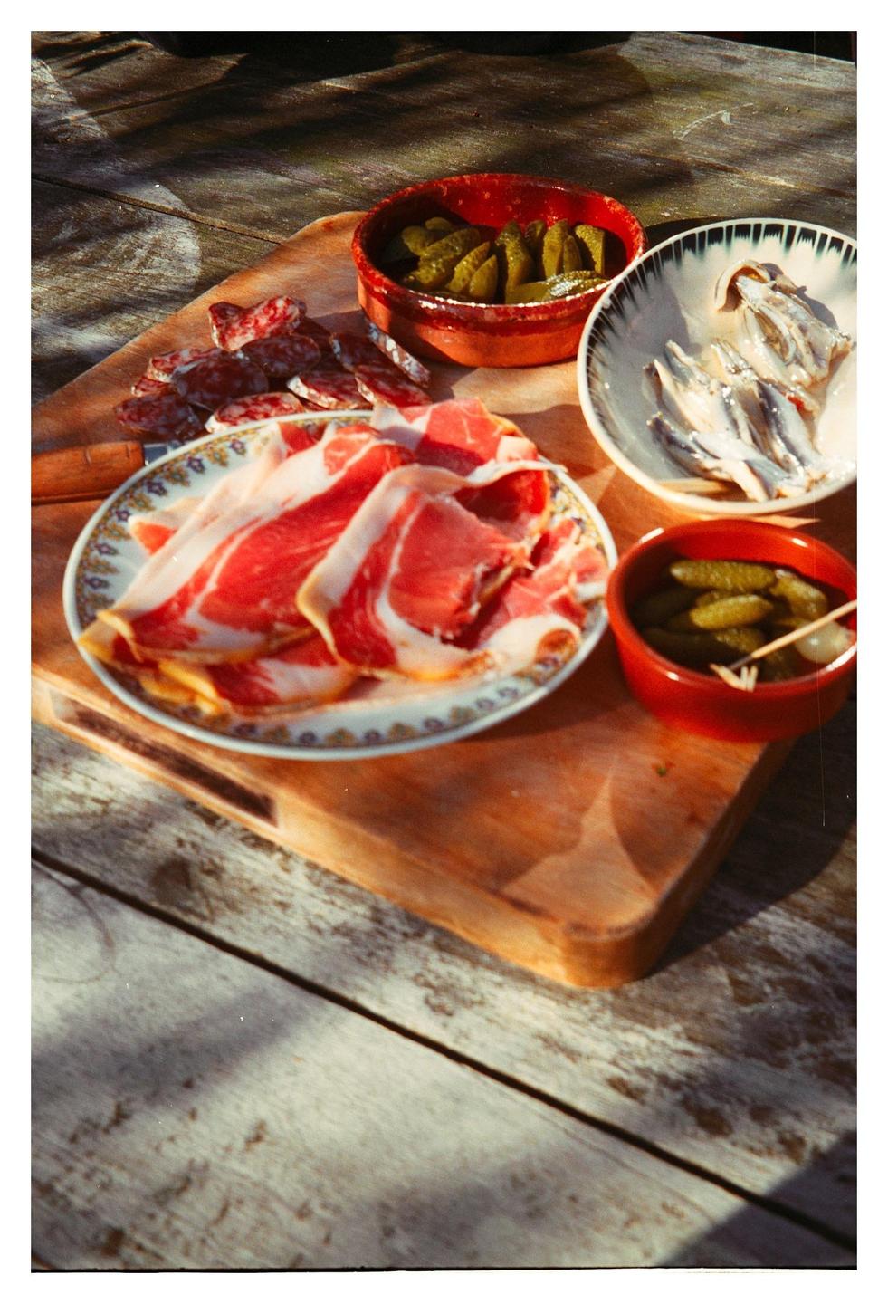 A wooden board holds plates of sliced meat, salami, anchovies, and pickles on a wooden table.