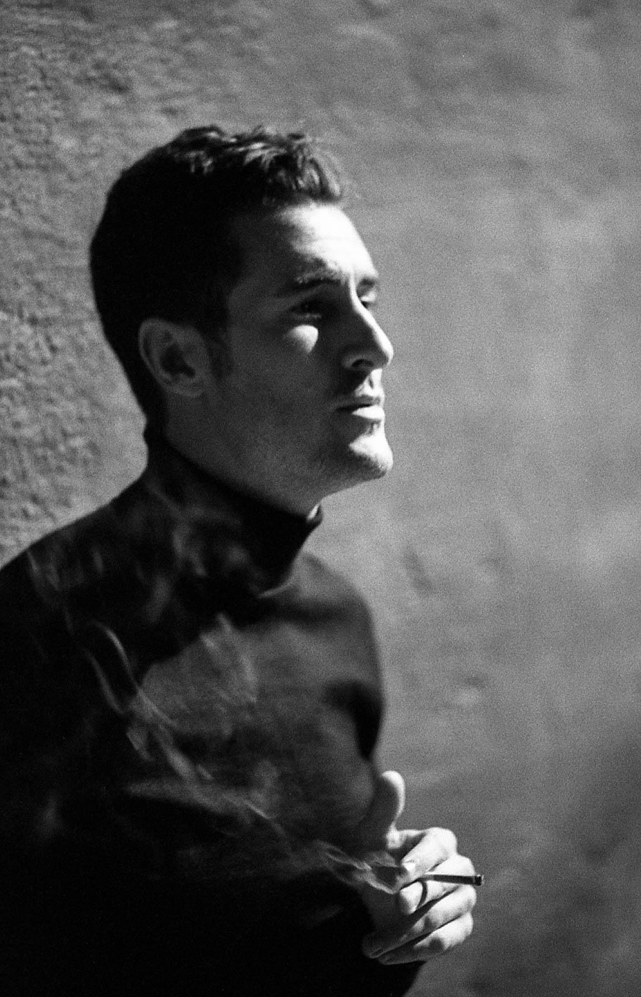 A man in a turtleneck sweater smokes a cigarette, with smoke rising around him.