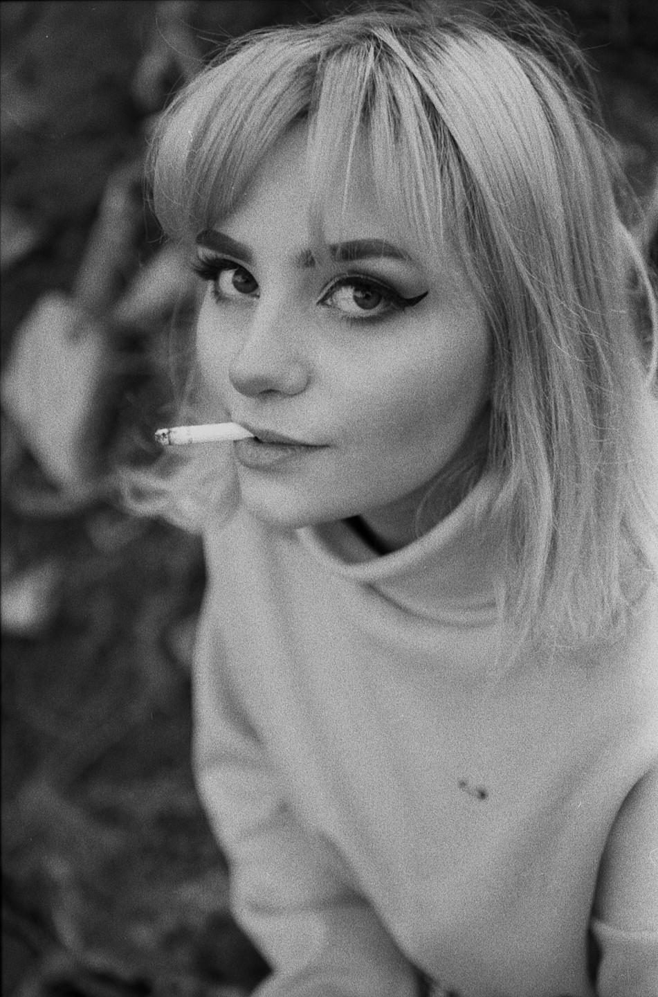 A woman with bangs and winged eyeliner looks up at the viewer while holding a cigarette in her mouth.