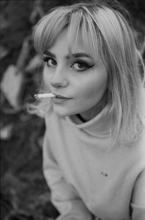 A woman with bangs and winged eyeliner looks up at the viewer while holding a cigarette in her mouth.