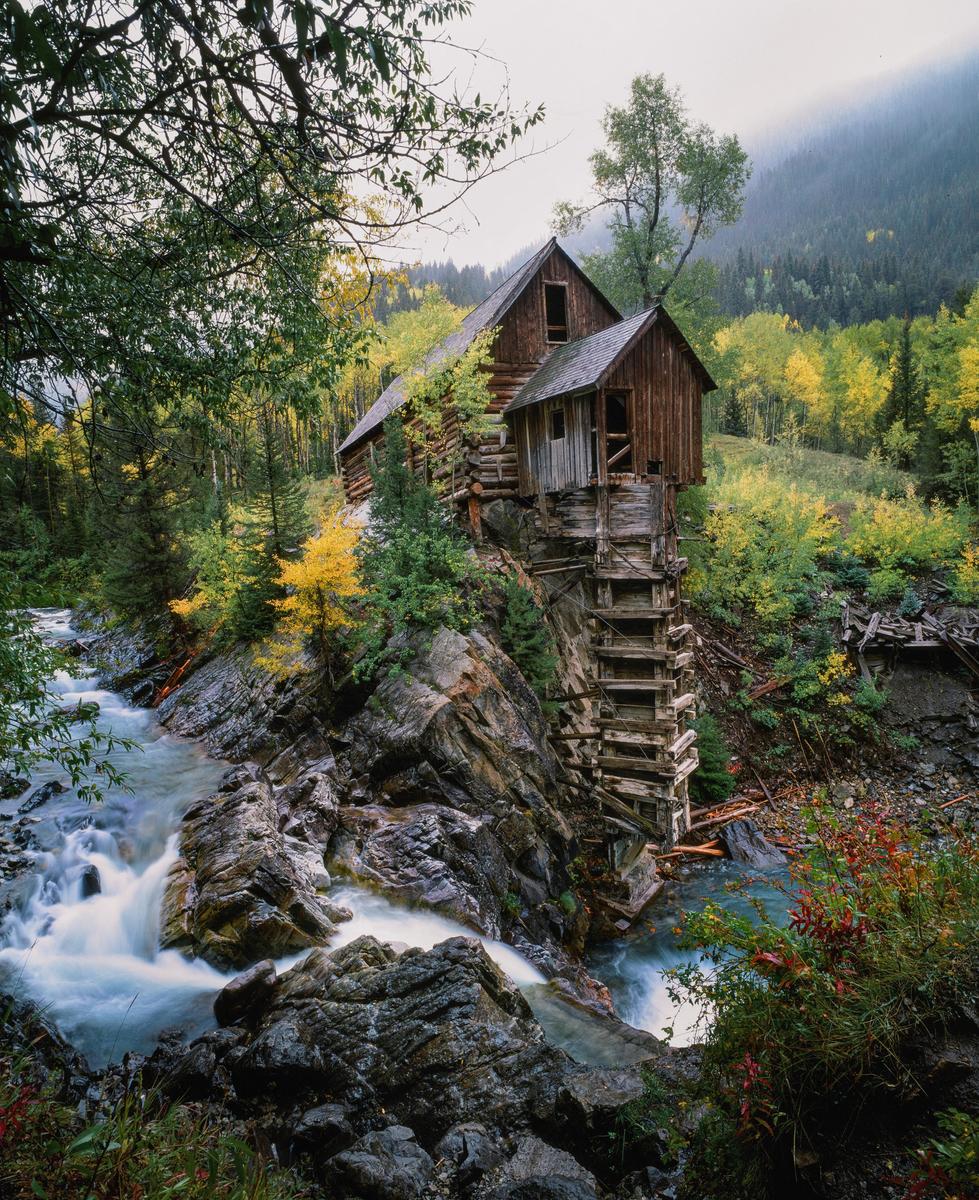 A wooden mill stands on a rocky cliff beside a rushing river, surrounded by autumn foliage.