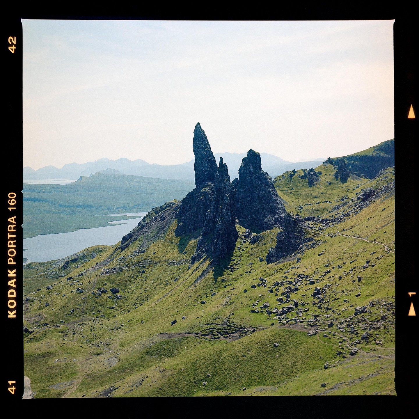 A large rock formation stands on a green hillside overlooking a body of water and distant mountains.
