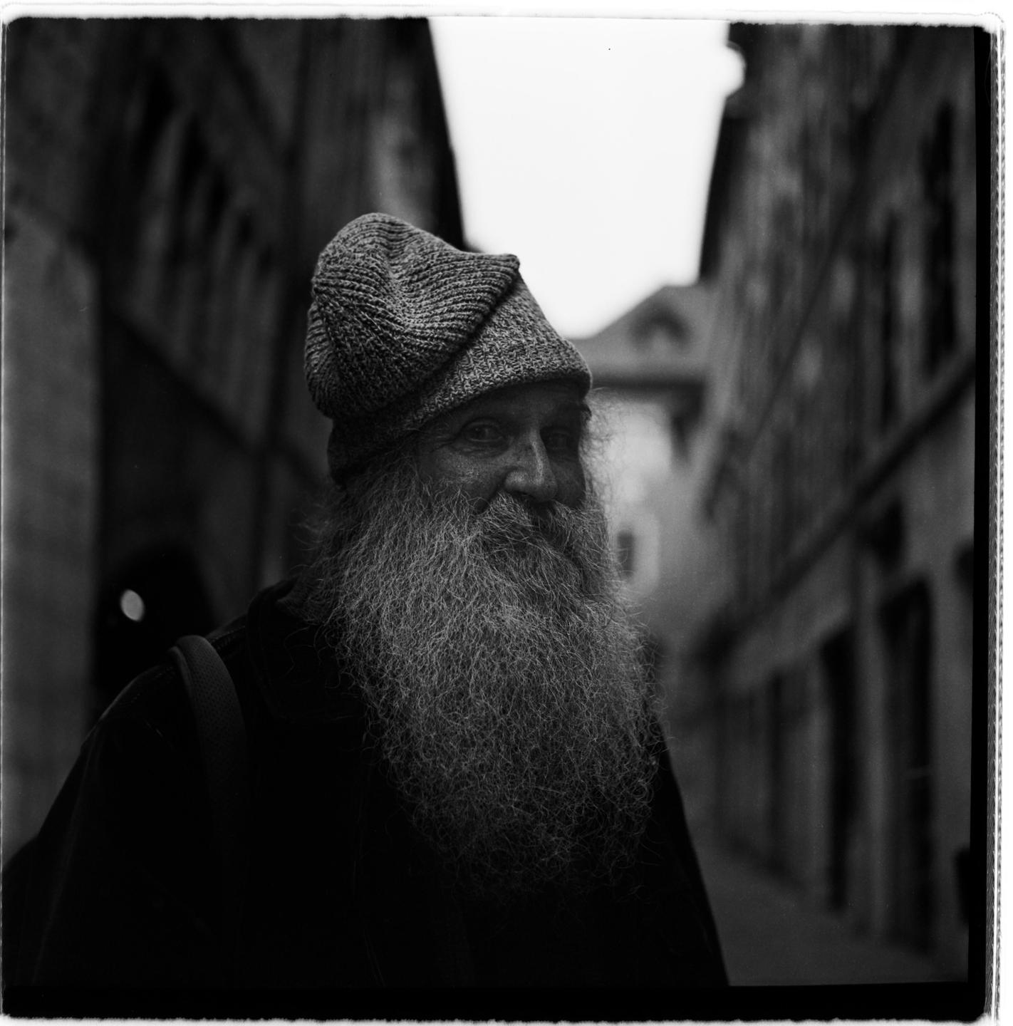 A man with a long beard and a knit hat smiles while standing in a narrow street.