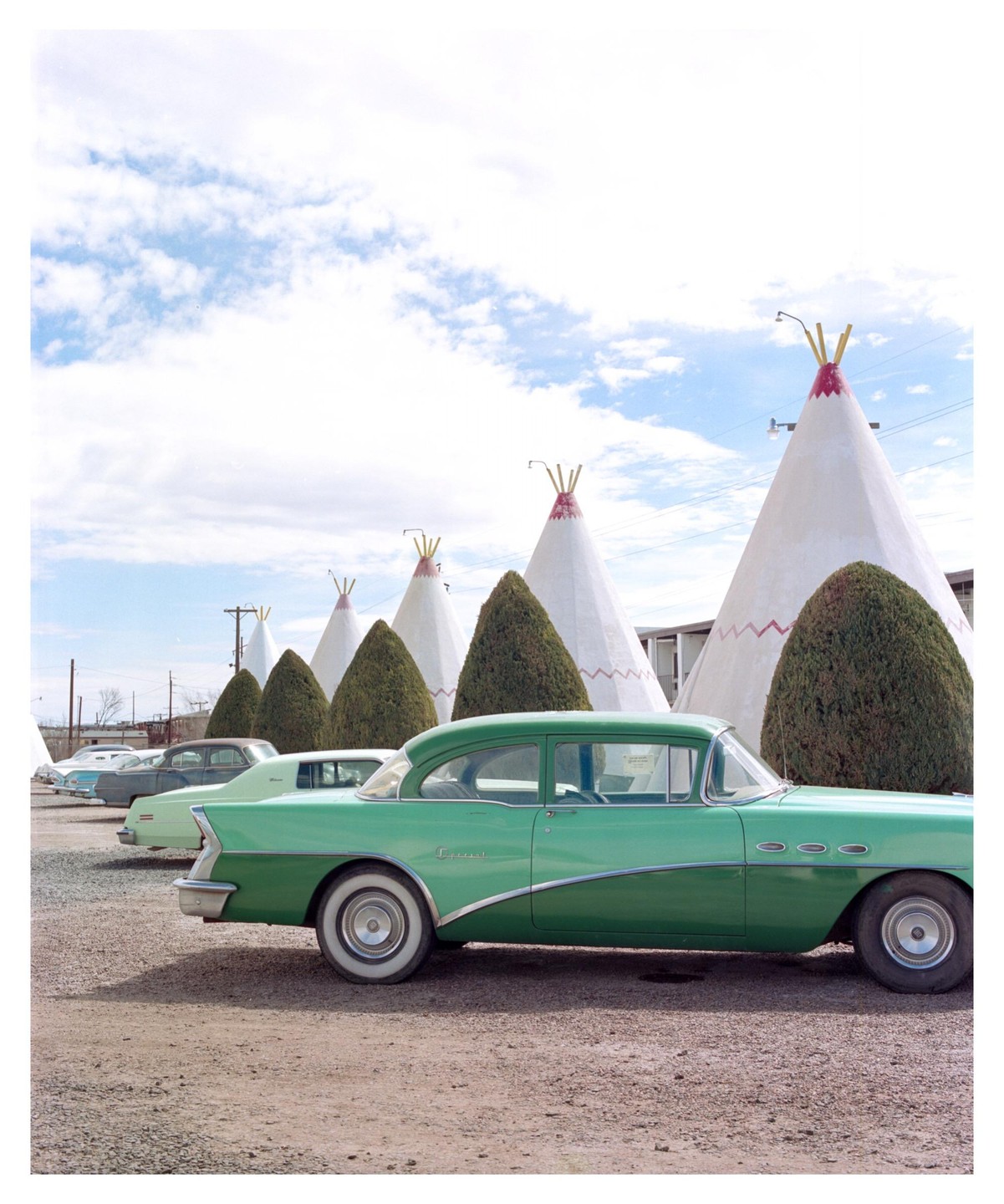 A green vintage car is parked in a gravel lot with several teepee-shaped buildings and manicured bushes in the background.
