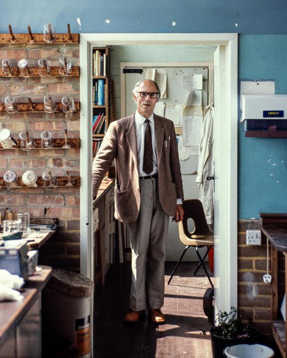 A man stands in a doorway, looking at the viewer, in a room with brick walls and laboratory equipment.