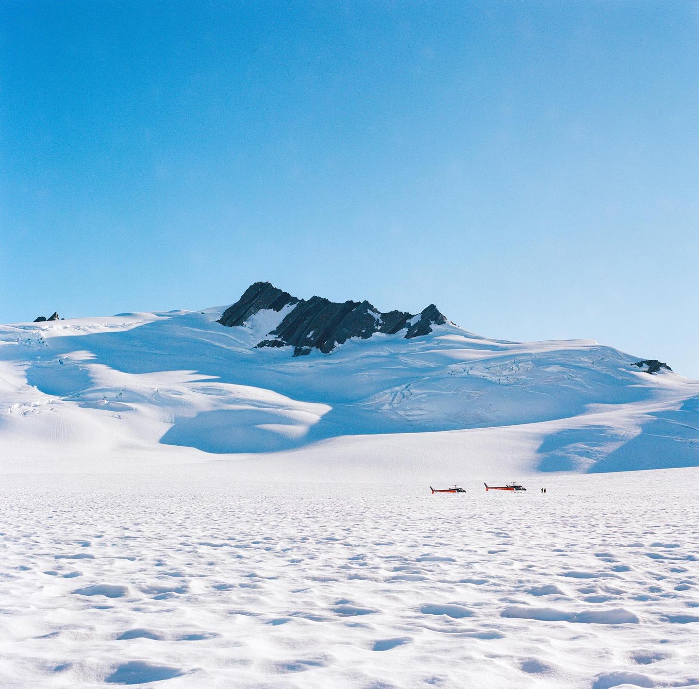 Two red helicopters are parked on a vast snowfield with a large snow-covered mountain in the background under a clear blue sky.