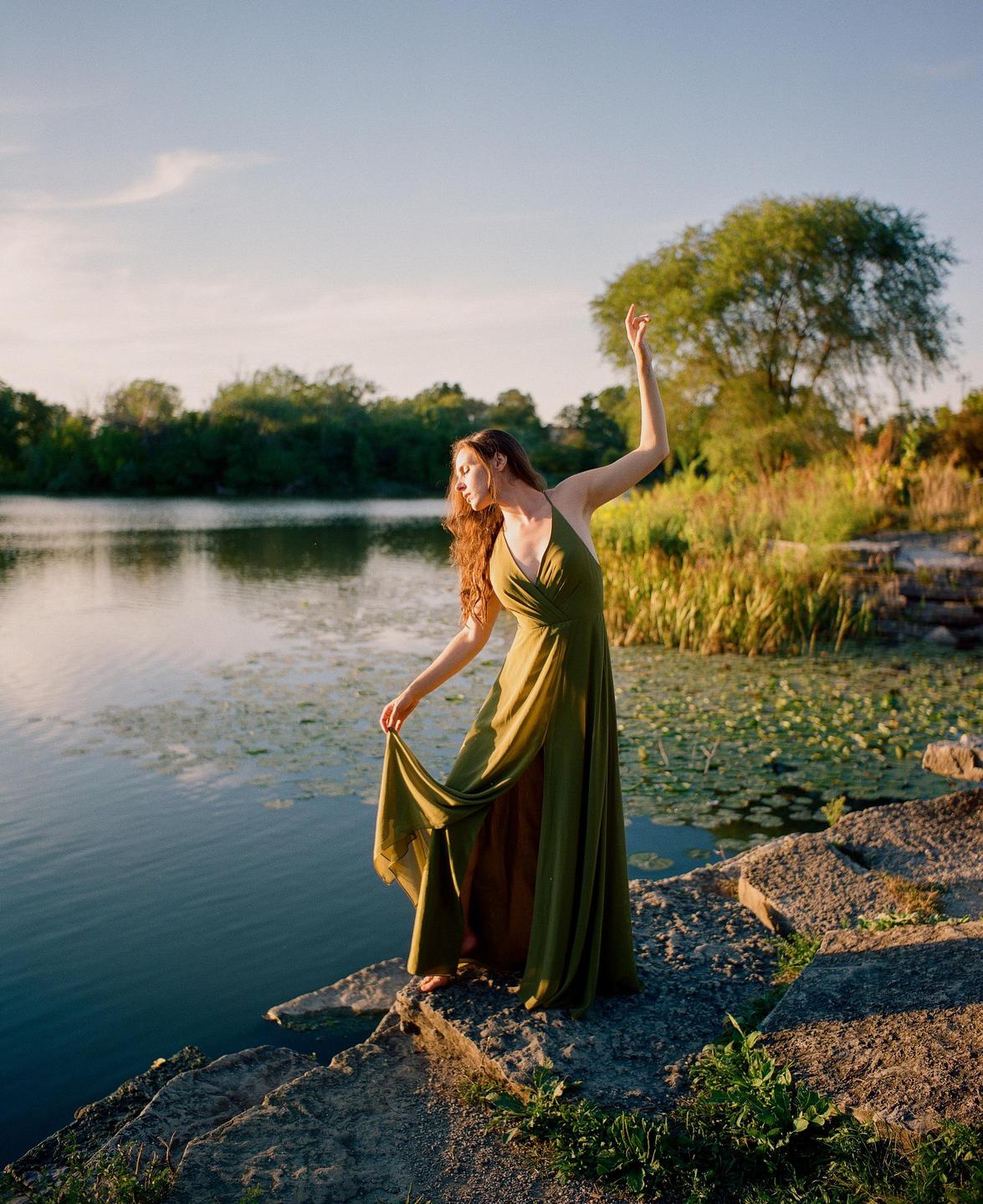 A woman in a green dress stands on rocks next to a lake with lily pads.