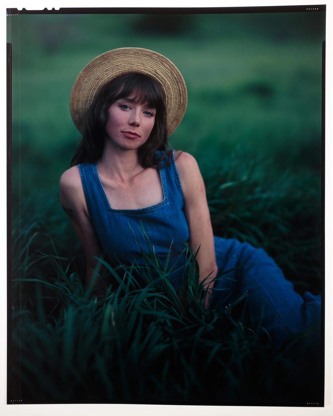A woman in a straw hat and denim dress sits in tall grass.