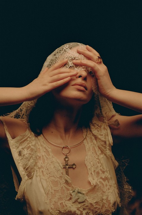 A woman with a lace veil covering her eyes wears a lace garment and a necklace with a cross pendant.