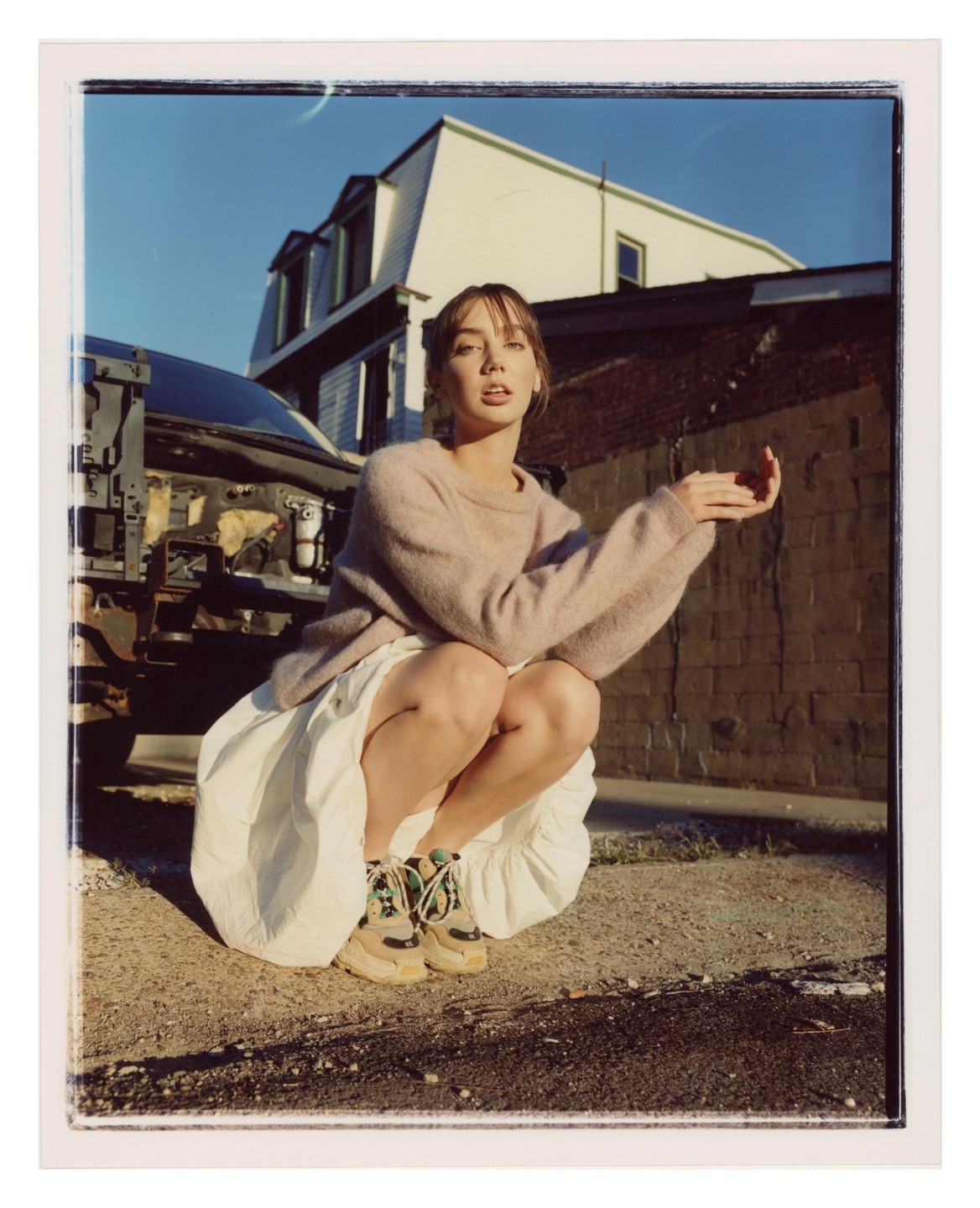 A woman squats on a paved surface next to a dismantled car, with a brick wall and a white building in the background.