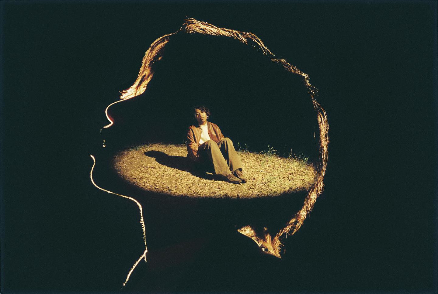 A woman sits in a spotlight on the ground, framed by a glowing silhouette of a head.