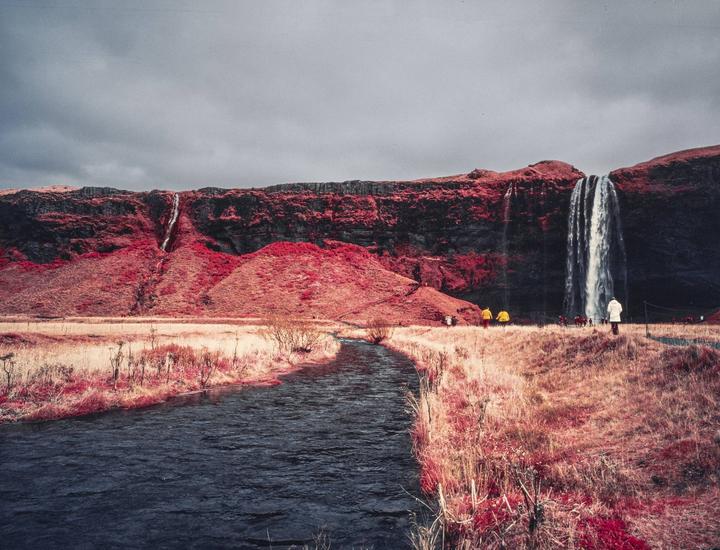 A river flows through a red landscape with a large waterfall cascading down a cliff under a cloudy sky.