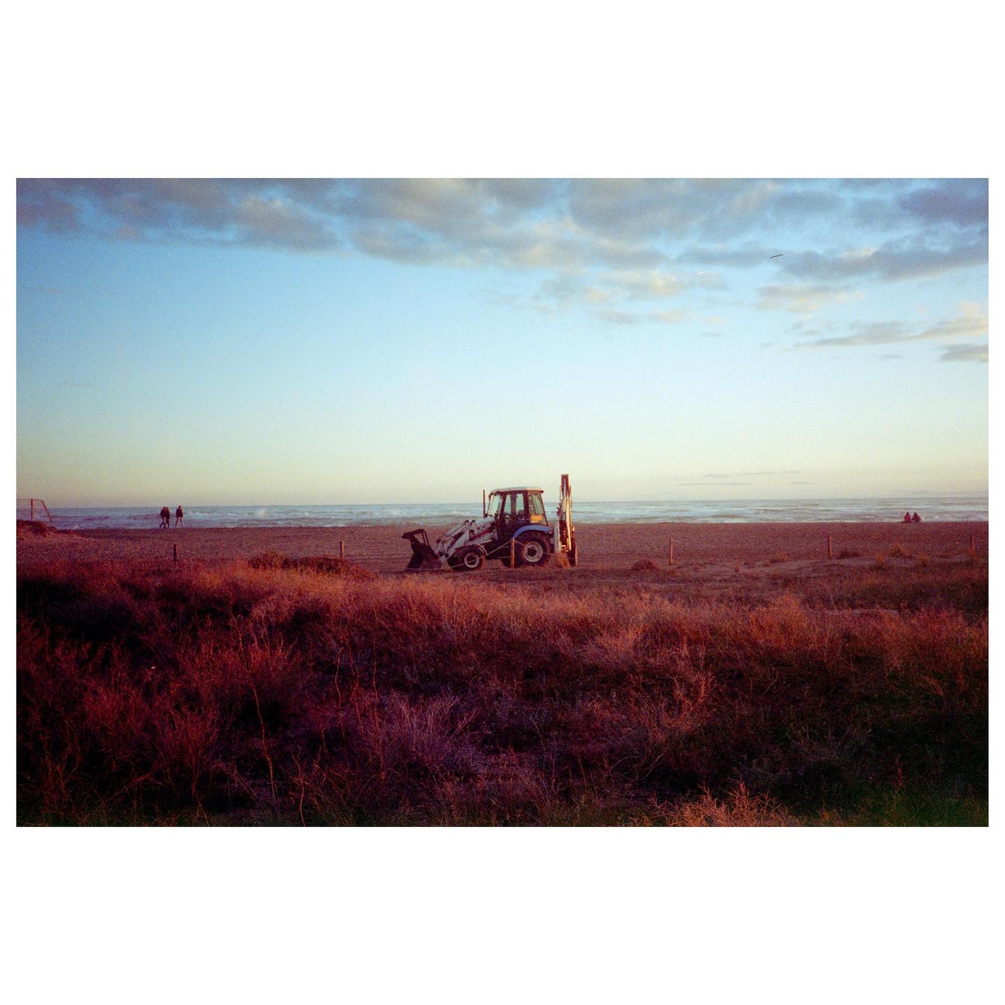 A blue and white tractor is parked on a beach with dry, reddish vegetation in the foreground and the ocean under a cloudy sky in the background.