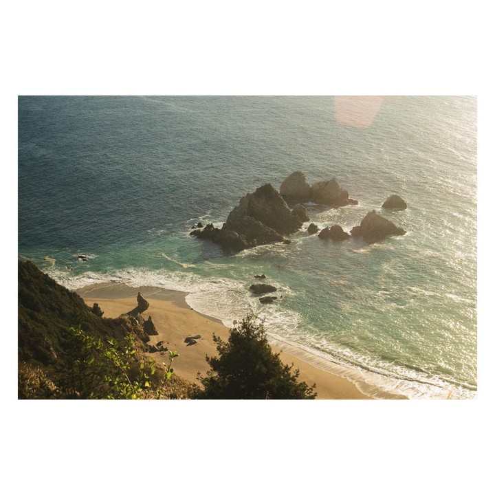 A high-angle view shows a sandy beach with waves crashing, several large rocks in the ocean, and a green hillside in the foreground.