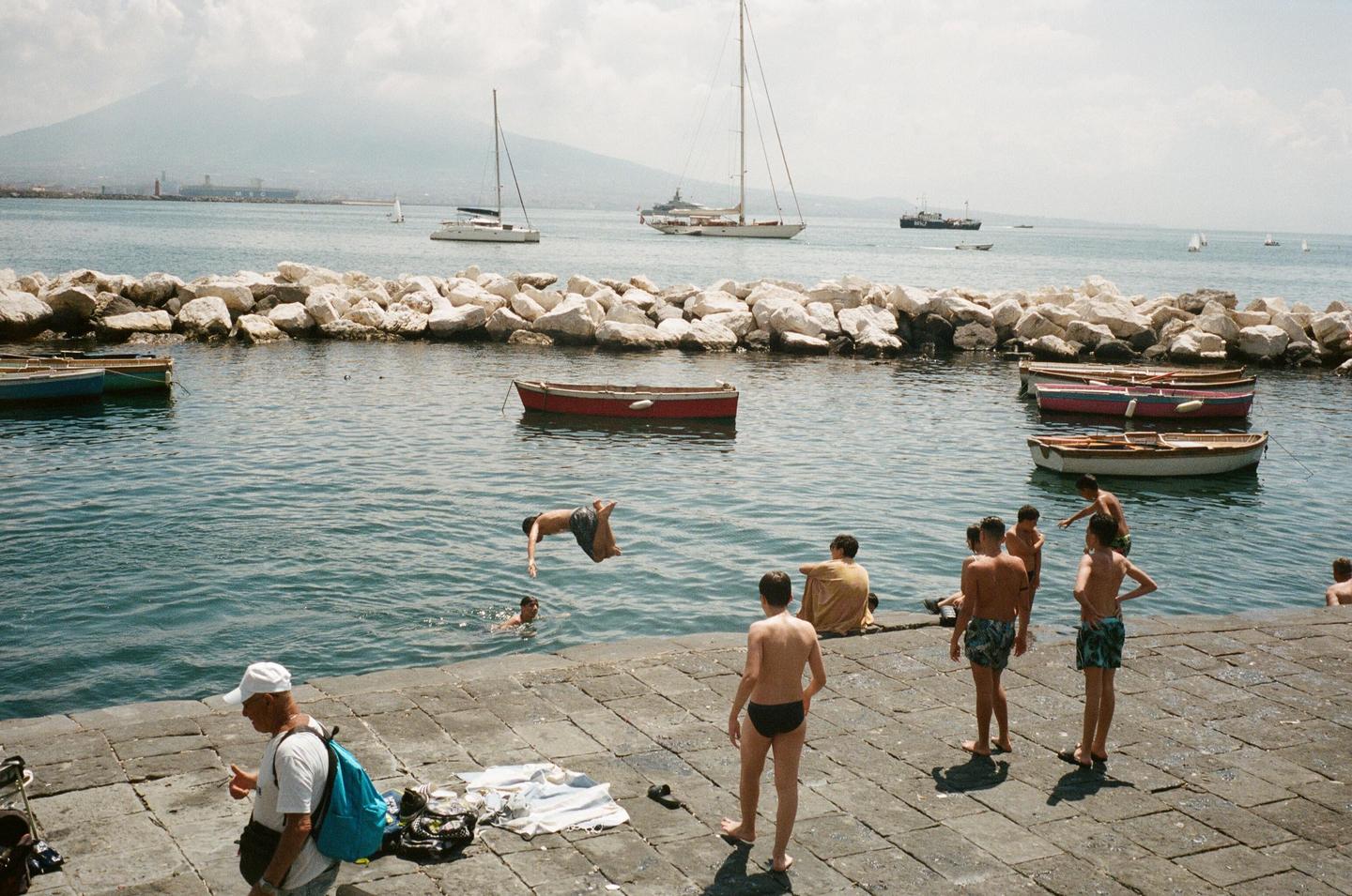 A boy dives into the water from a stone pier where other boys are gathered, with boats and a mountain in the background.