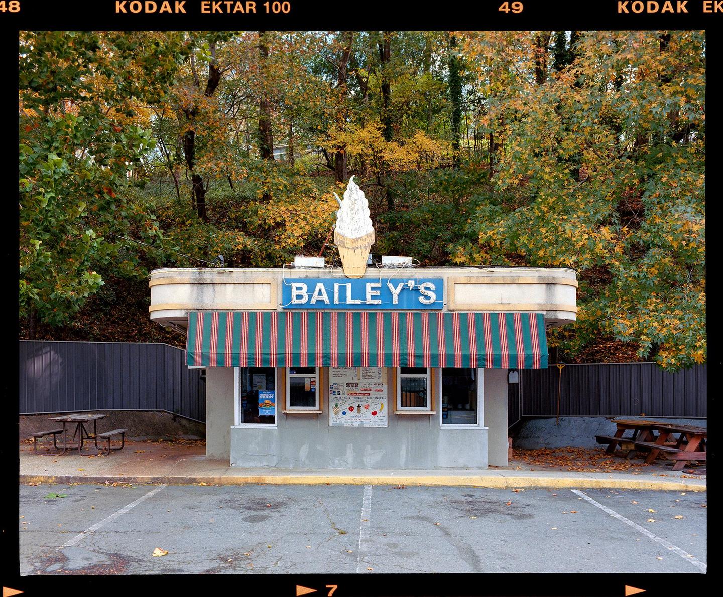An ice cream stand with a large ice cream cone sign on its roof is surrounded by trees with autumn foliage.