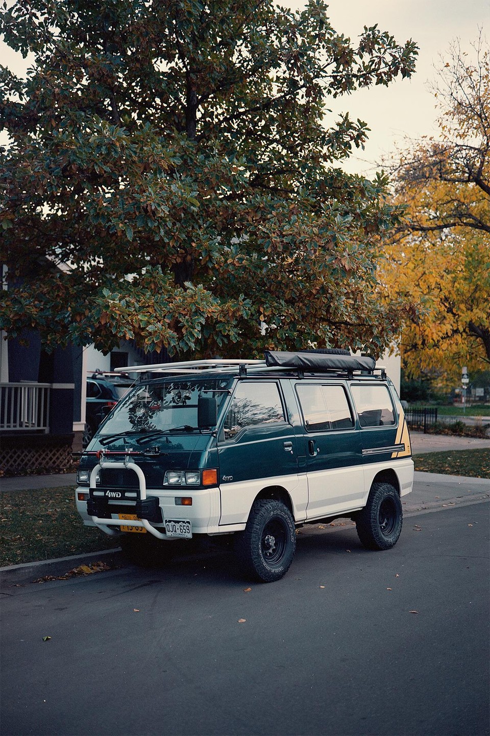 A two-tone van with a roof rack is parked on a street next to a large tree with autumn foliage.