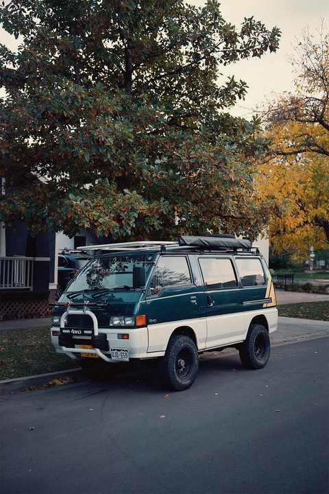 A two-tone van with a roof rack is parked on a street next to a large tree with autumn foliage.