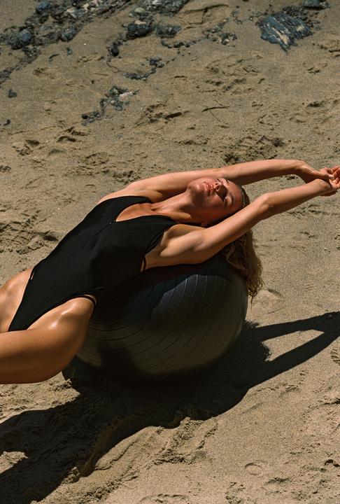 A woman in a black swimsuit stretches on an exercise ball on a sandy beach.