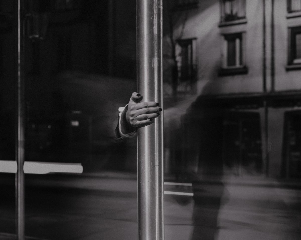 A hand with dark nail polish holds a metal pole in front of a reflective surface with building reflections.