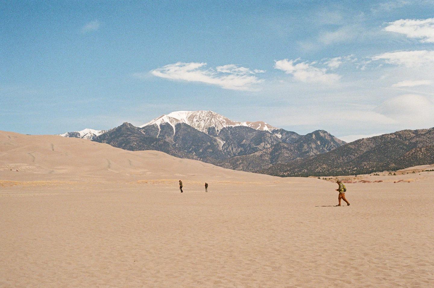 Three people walk across a vast desert landscape with sand dunes and snow-capped mountains under a blue sky.