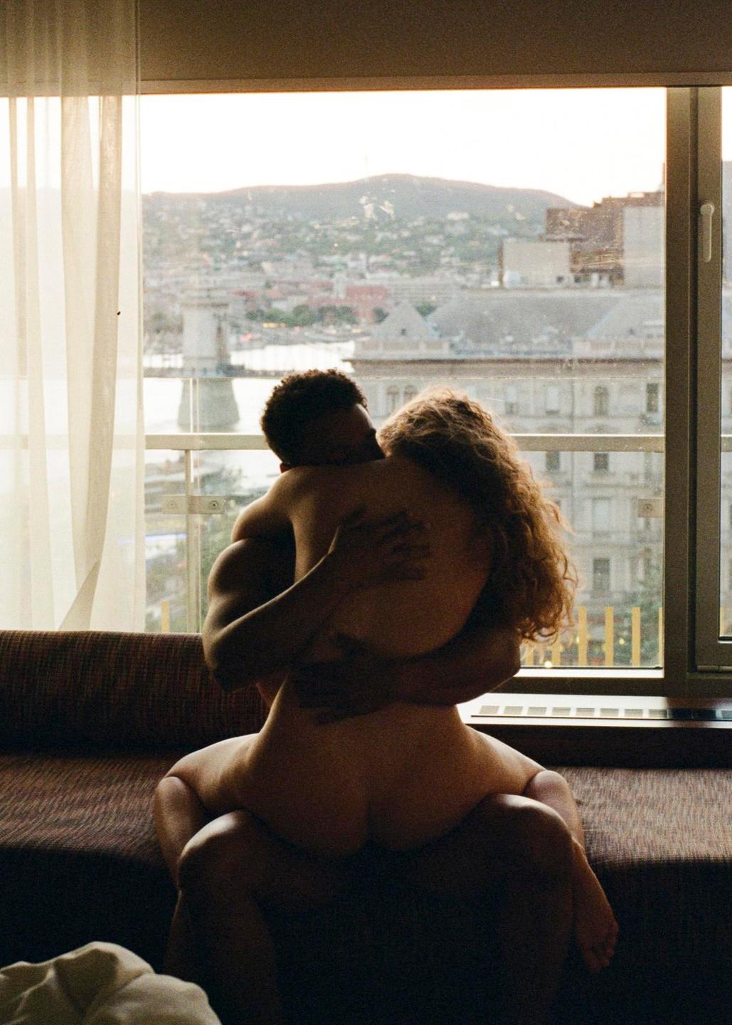 A couple embraces in front of a window overlooking a city and mountains.