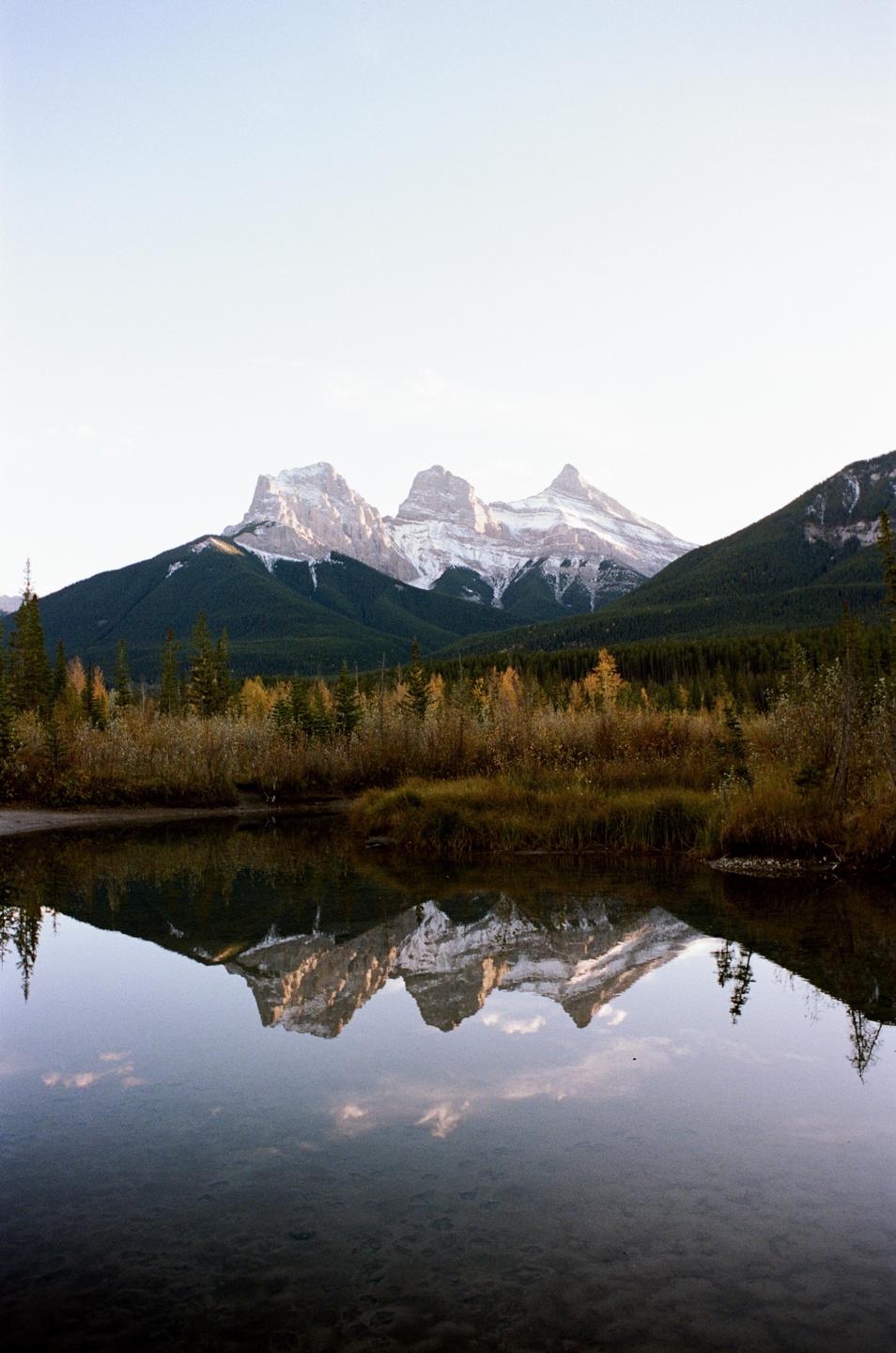 Snow-capped mountains are reflected in a calm body of water, surrounded by autumn foliage and evergreen trees.