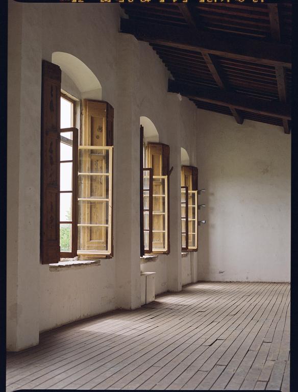 Sunlight streams through a row of open windows with wooden shutters onto a wooden floor.