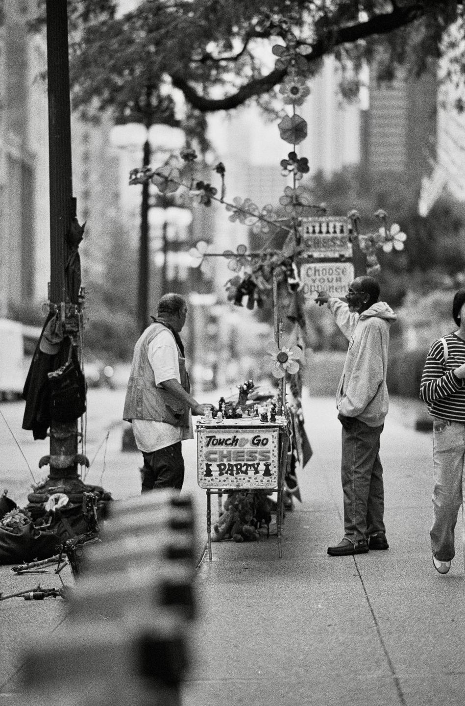 Two men are standing on a city street next to a chess game stand, with one man pointing towards something out of frame.