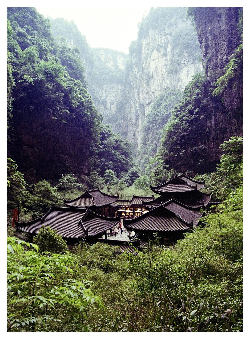 A cluster of traditional buildings with dark tiled roofs is nestled in a lush green valley surrounded by towering cliffs.